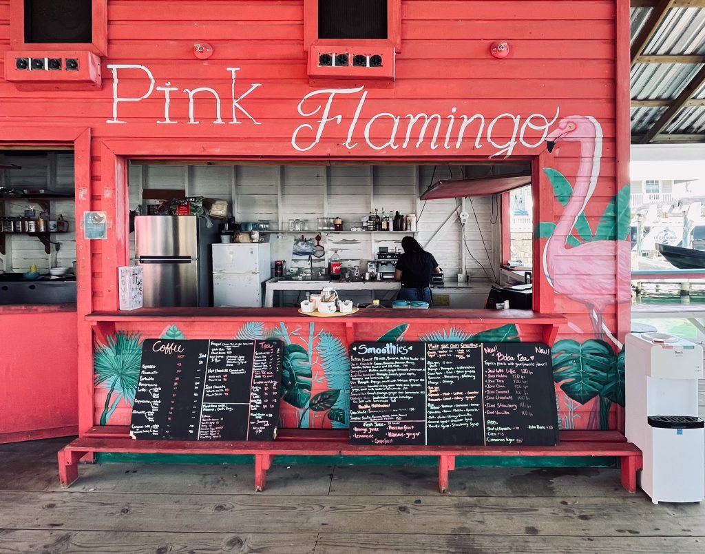 View of a pink shack restaurant, "Pink Flamingo", on a wooden deck. A woman is working in the kitchen with her back turned. Two slate menu boards are lined up along the lower edge of the counter. Part of a marina is visible in the background in the top right corner.
