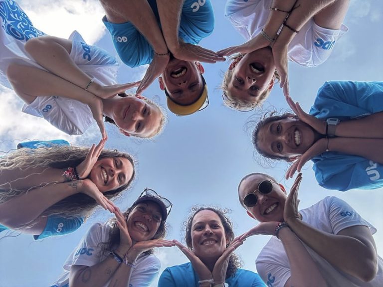 low angle view, eight people in a circle, alternatively wearing blue or white shirts, holding their hands under their chin to form a star pattern.